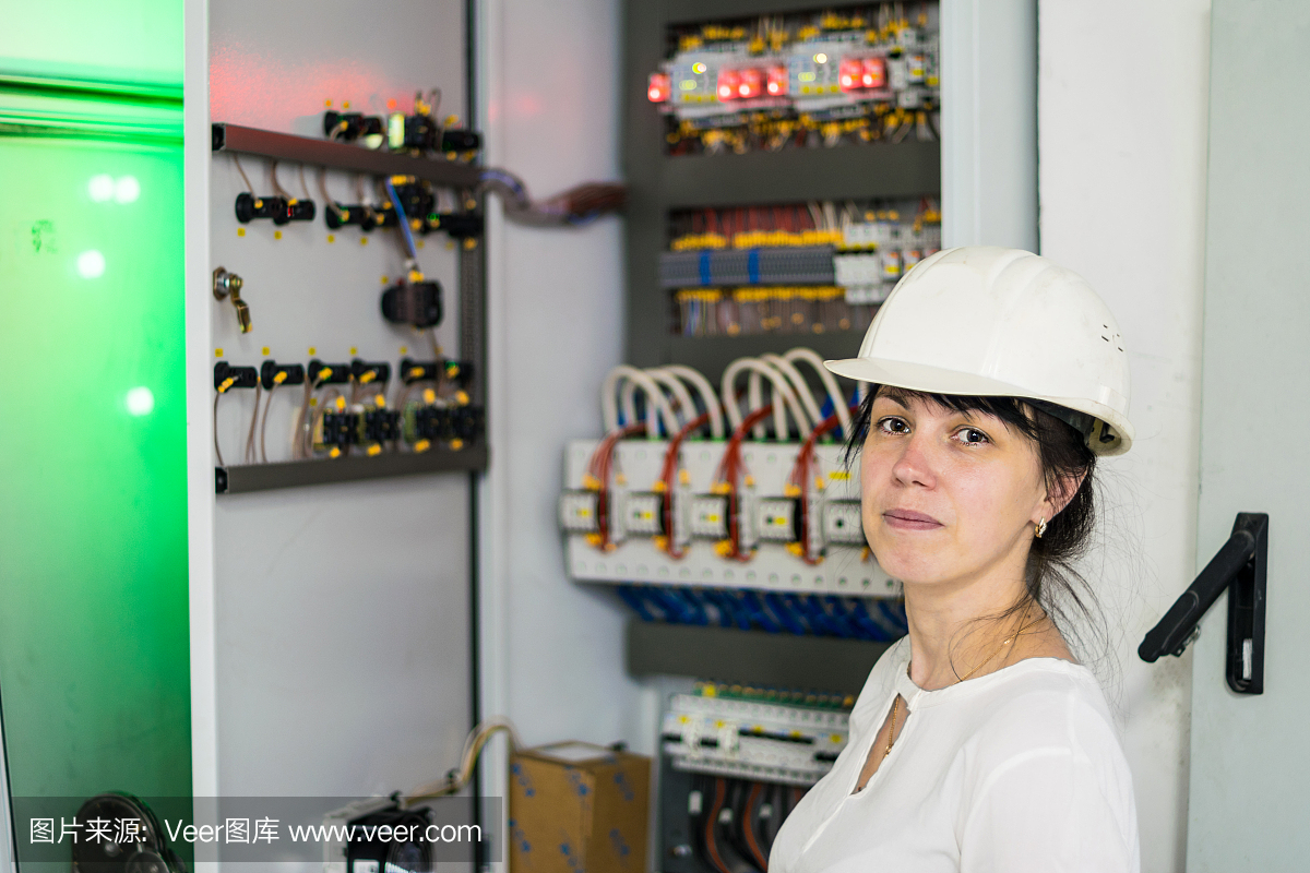 一名戴著白色頭盔的電工婦女的肖像,旁邊是高壓電氣設備。一個戴著工程頭盔的女孩站在保險絲盒的背景上。